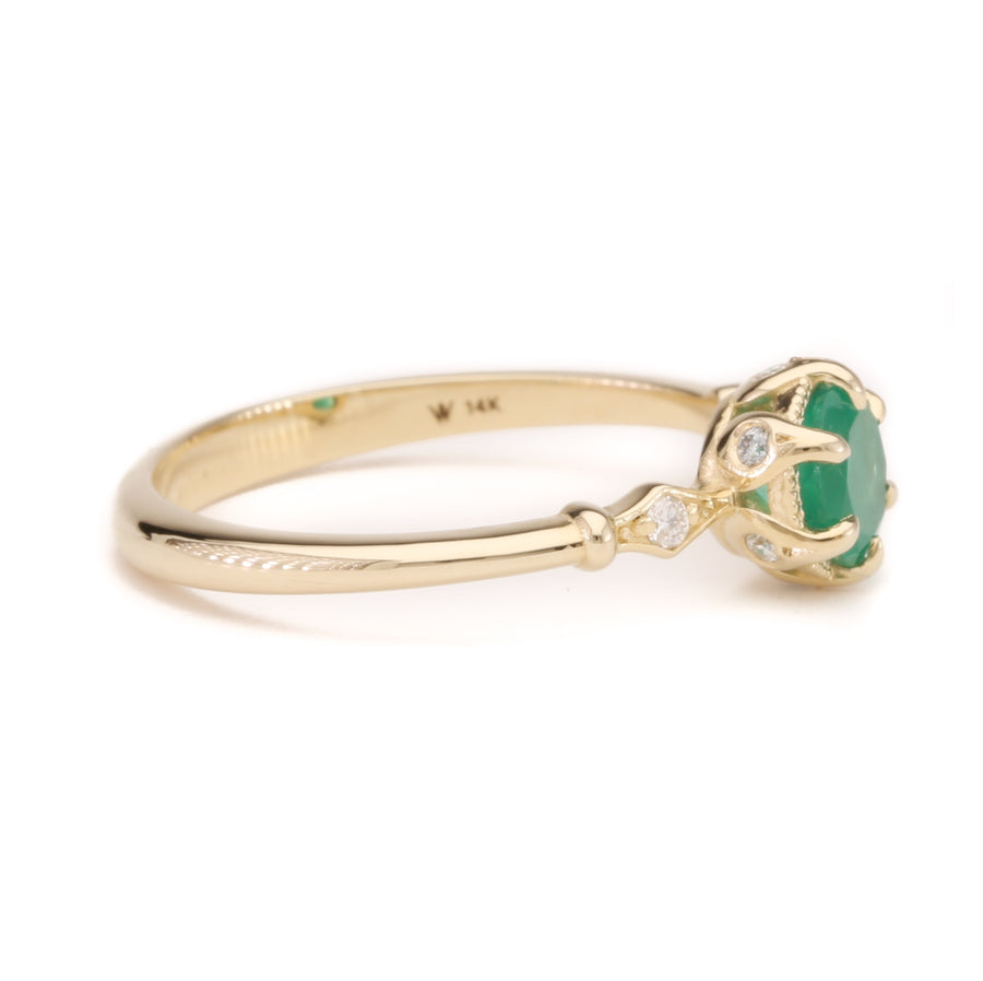 A close-up image of a 14K solid yellow gold ring featuring a vibrant green emerald gemstone in a fairytale-inspired setting, displayed on a white background. The ring has a delicate, elegant band with intricate detailing around the emerald.