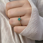 A close-up image of a hand wearing a delicate 14K solid yellow gold ring featuring a round green emerald gemstone in a simple, elegant setting. The ring is showcased on the finger, highlighting its fine craftsmanship and vibrant emerald centerpiece.