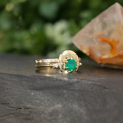A close-up image of two elegant 14kt solid gold rings: one features a round emerald gemstone in a classic prong setting, while the other is a crescent horn-shaped band. Both rings are displayed side by side on a neutral background, highlighting their fine craftsmanship and vibrant green gemstone.
